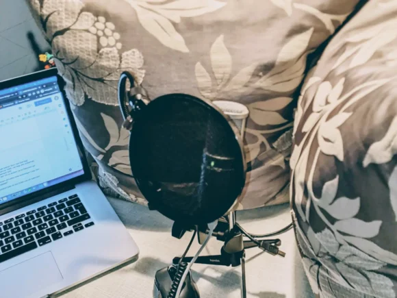 Aufnahme- oder Podcasting-Setup. Zu sehen ist ein Laptop mit einem geöffneten Dokument auf dem Bildschirm, ein Mikrofon mit einem Pop-Filter sowie einige Kissen im Hintergrund.